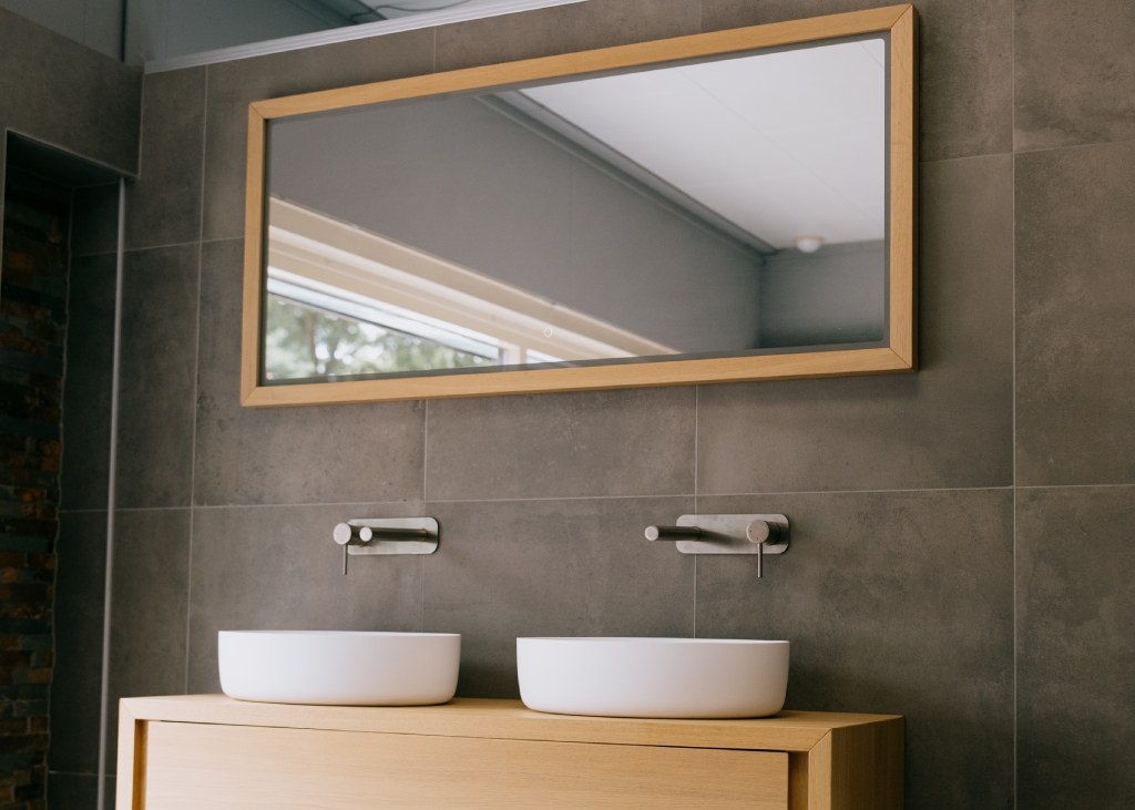 Wilt u een badkamer monteren in Zuidwolde? Wij ontzorgen u volledig. Een langwerpig houten wasmeubel met twee witte kommen en een rechthoekige spiegel erboven.