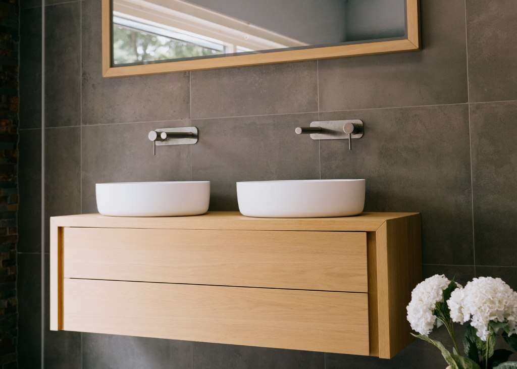 Een langwerpig houten wasmeubel met twee witte kommen en een rechthoekige spiegel erboven